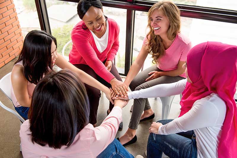 Group of diverse women put their hands together in stack empowering each other in breast cancer awareness campaign meeting